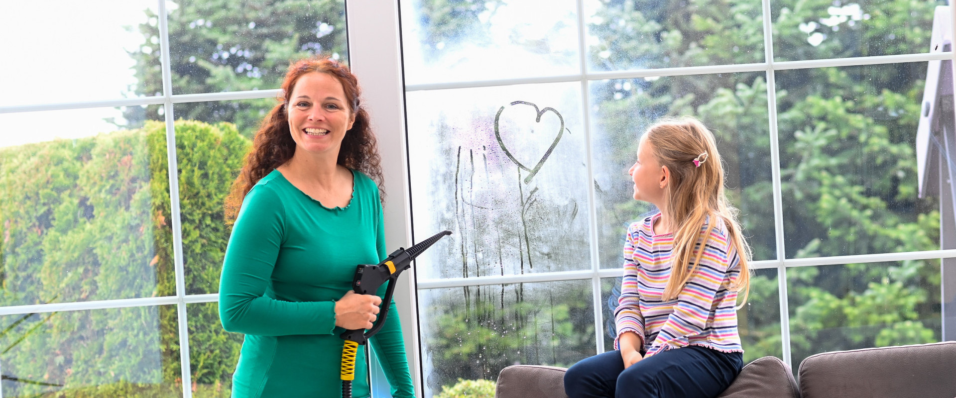 A little girl draws a heart with her finger on the window pane, which her mother is cleaning with dry steam
